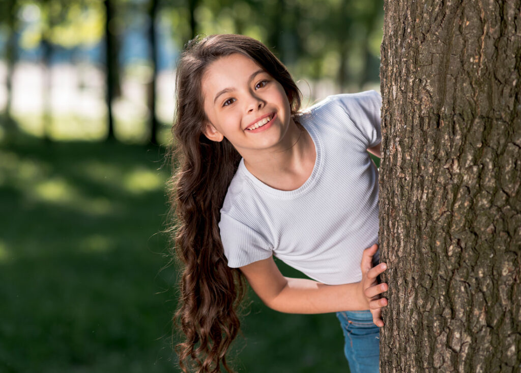 smiling cute girl looking out from tree trunk outdoors1 2
